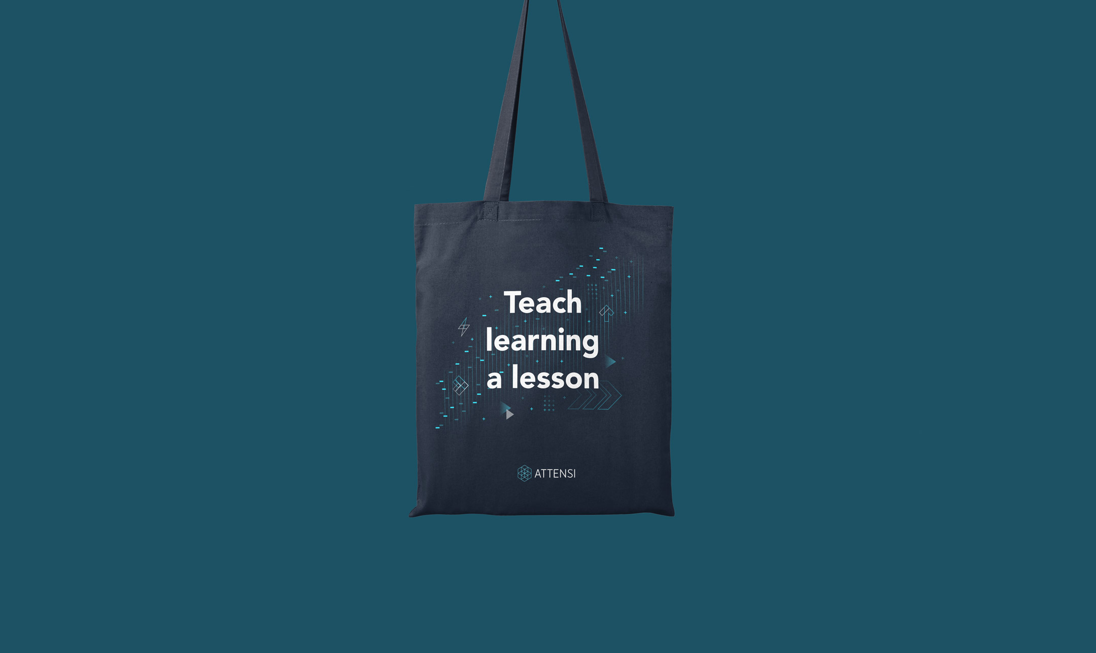 The image displays a dark tote bag against a teal blue background. On the tote bag, there is white and bright blue text that reads "Teach learning a lesson" alongside abstract digital designs, including geometric shapes and circuit-like patterns. The company logo "ATTENSI" is also visible at the bottom of the design. The overall style is modern and tech-oriented.