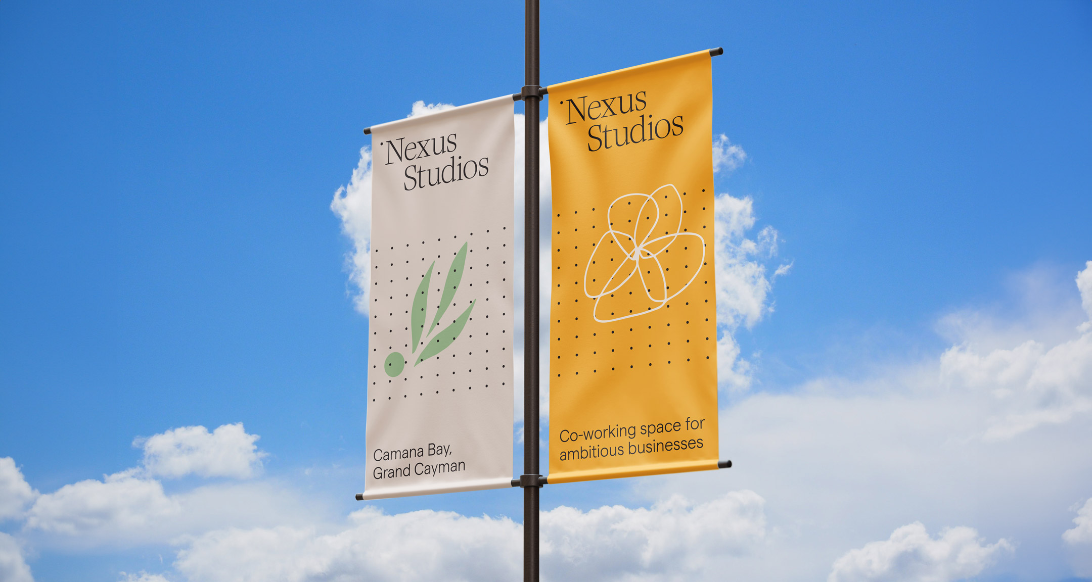 Two vertical banners mounted on a single flagpole. The left banner is white with the logo of "Nexus Studios," featuring a stylised green leaf and black dot pattern along with the text "Camana Bay, Grand Cayman" in a serif font. The right banner is golden yellow with a large, abstract, white outline of flower drawing that similar to an atom model, surrounded by a dotted pattern. It includes the text "Nexus Studios" at the top and "Co-working space for ambitious businesses" at the bottom, both in a serif font. The banners are depicted fluttering slightly, suggesting a gentle breeze.