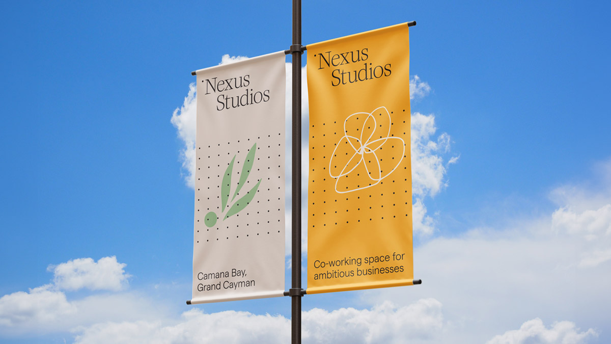 Two vertical banners mounted on a single flagpole. The left banner is white with the logo of "Nexus Studios," featuring a stylised green leaf and black dot pattern along with the text "Camana Bay, Grand Cayman" in a serif font. The right banner is golden yellow with a large, abstract, white outline of flower drawing that similar to an atom model, surrounded by a dotted pattern. It includes the text "Nexus Studios" at the top and "Co-working space for ambitious businesses" at the bottom, both in a serif font. The banners are depicted fluttering slightly, suggesting a gentle breeze.