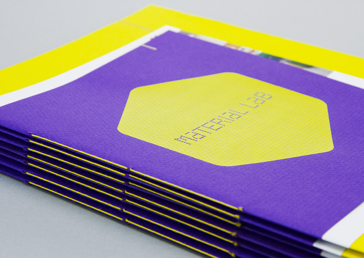 A stack of 7 Material Lab 20 page booklets. The cover stock is Colorplan Matrix emboss, in purple. On the cover is a large yellow screen printed hexagon with the Material Lab logo inverted in the centre. The sides of the booklet are visible and are saddle stitched with yellow thread.