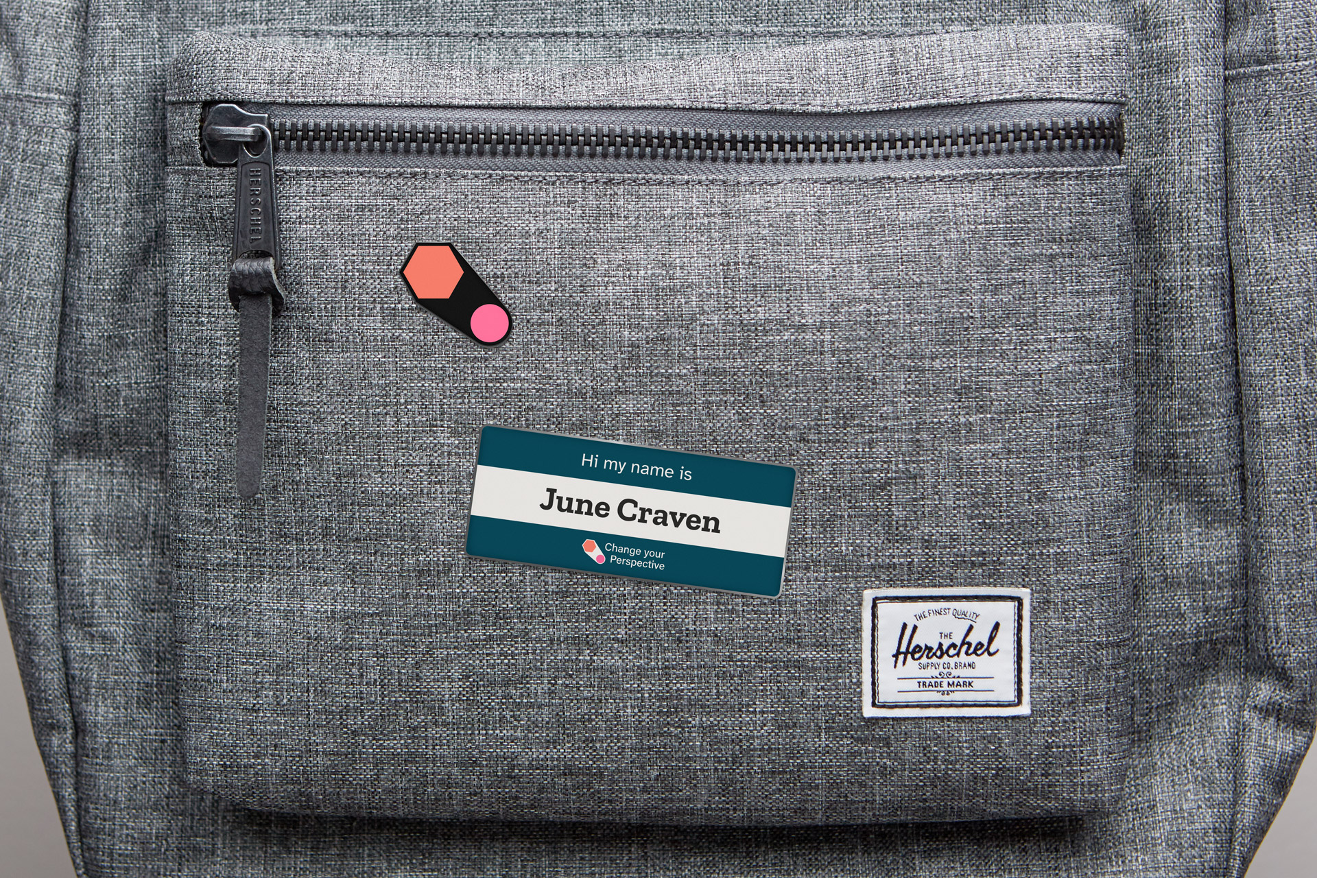 A grey backpack with a Change Your Perspective brand logo enamel pin and a name tag reading 'Hi my name is June Craven' attached to the front pocket.